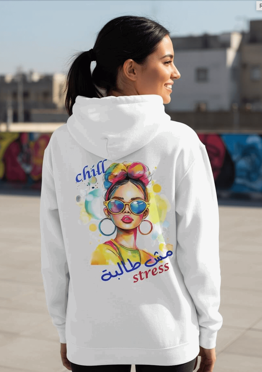 Woman wearing a white hoodie with colorful graphic and Arabic text in an urban setting. Hoodie says Chill, no stress.
