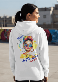 Woman wearing a white hoodie with colorful graphic and Arabic text in an urban setting. Hoodie says Chill, no stress.